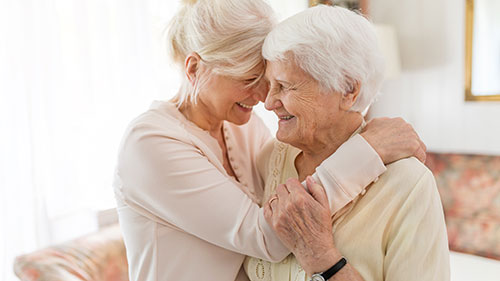 Two elderly women standing and hugging in the middle of a room